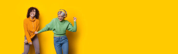 Two cheerful curly haired sisters are listening to music and smile on a yellow studio wall with free space using headphones