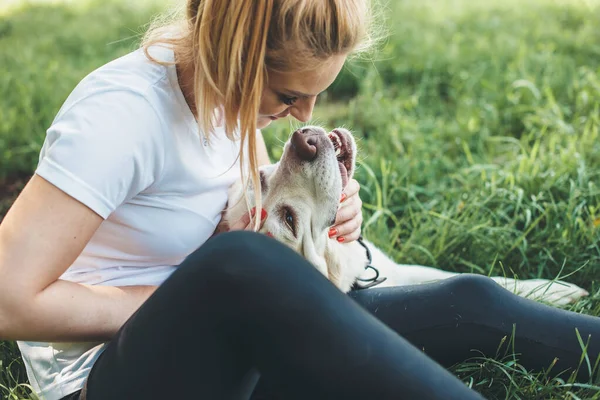 Side view photo of a blonde woman in casual clothes playing on the grass with her labrador embracing and kissing it — Stock Photo, Image