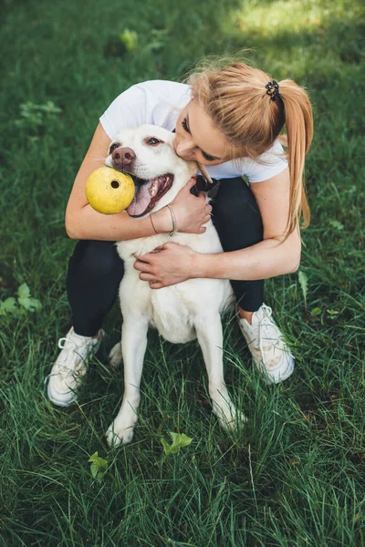 Szőke kaukázusi nő átöleli a kutyáját, miközben egy zöld parkban játszik egy labdával. — Stock Fotó