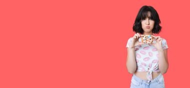 Kafkasyalı bir kadın, kırmızı bir stüdyo duvarında tatlı bir çörek tutarken komik bir yüz ifadesi takınıyor.