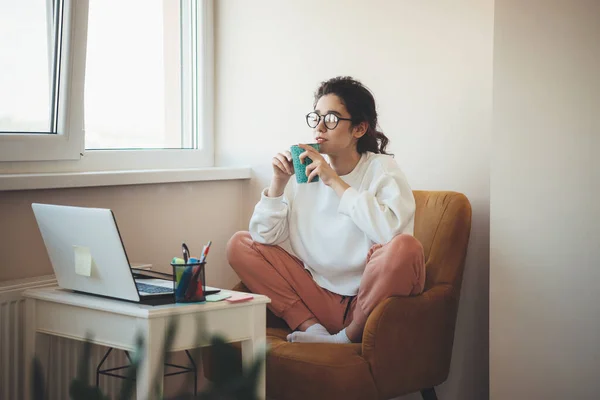 Foto de una mujer concentrada con vasos bebiendo té en casa sentada en un sillón y viendo algo en el portátil — Foto de Stock
