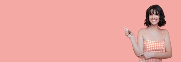 Жінка в літньому одязі вказує на рожеву стіну студії з вільним простором, посміхаючись у камеру. — стокове фото