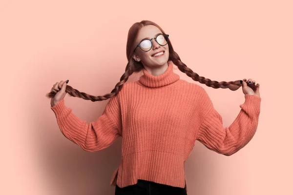 Mujer infantil con pecas y el pelo rojo está mirando a través de gafas en la cámara y sonreír en una pared de estudio rosa tocando su cabello — Foto de Stock