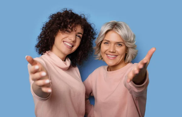 Foto de cerca de dos mujeres con el pelo rizado sonriendo a la cámara y un gesto atractivo con palmas en una pared de estudio azul — Foto de Stock