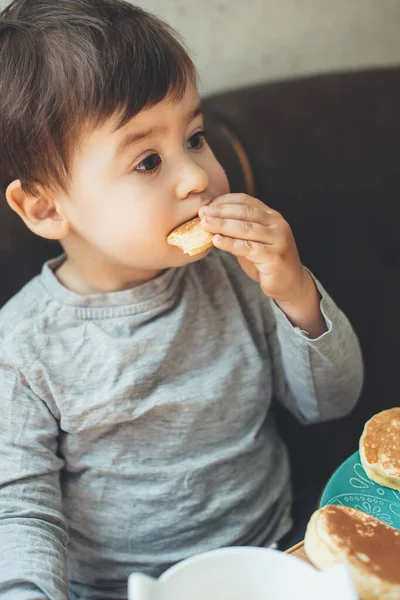 Menino pequeno está comendo panquecas na mesa enquanto olha para alguém — Fotografia de Stock