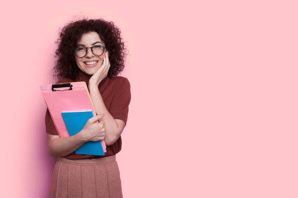 Surprised curly haired student is touching her cheek smiling on a pink wall with free space holding some folders