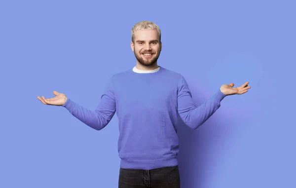 Foto monocromática de um homem barbudo com cabelo loiro comparando duas coisas em suas palmas sorrindo em uma parede de estúdio azul — Fotografia de Stock