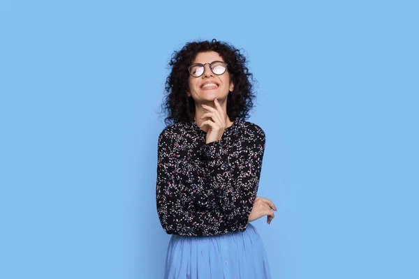 Adorabile donna con i capelli ricci e occhiali sta sorridendo alla fotocamera su una parete blu studio — Foto Stock