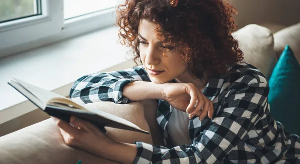 Soustředěná žena s kudrnatými vlasy čte knihu, zatímco sedí v posteli u okna — Stock fotografie