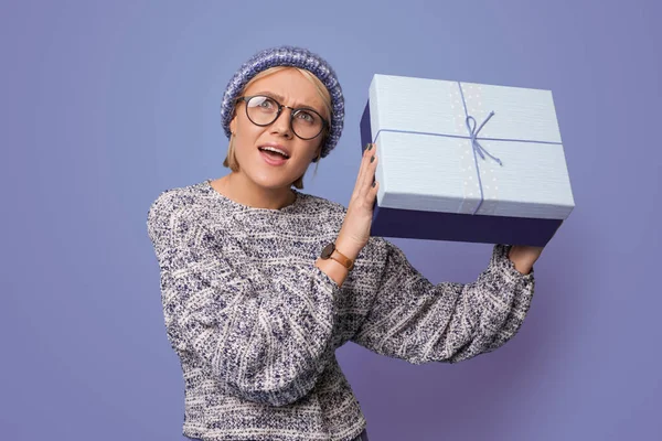 Mujer rubia posando sorprendida mientras agita un regalo en una pared de estudio azul con gafas y sombrero — Foto de Stock