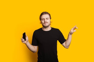 Uzun saçlı, sakallı, siyah tişörtlü, telefon ve kulaklıkla derin derin düşünen beyaz bir adam.