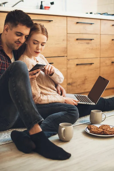 Vidám pár ül a padlón a konyhában egy laptop és telefon használata közben teát kortyolgat süteményekkel reggel — Stock Fotó