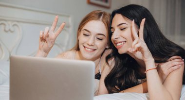 Kızıl ve siyah saçlı beyaz kız kardeşler, bilgisayar kullanarak yatakta bir video görüşmesi yaparken gülümsüyor ve işaret ediyorlar.