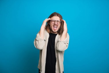 Stresli, beyaz, uzun kahverengi saçlı ve gözlüklü bir adam mavi bir duvarda baş ağrısı işareti yapıyor.