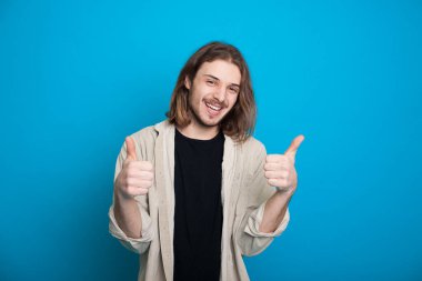 Uzun saçlı ve sakallı mutlu beyaz adam mavi bir duvarda işaret ediyor.