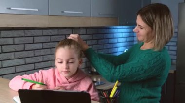 Kafkasyalı anne oturma odasındaki dijital tablete bakıp çizerken kızının saçını bağlıyor.