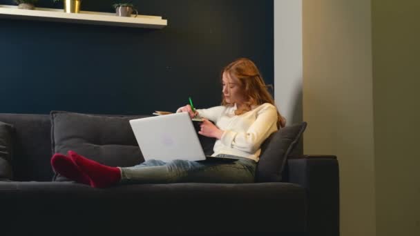 주근깨가 있는 백인 생강 여성이 컴퓨터로 침대에 누워 온라인 강의를 듣는 동안 몇 가지 음을 적고 있습니다. — 비디오