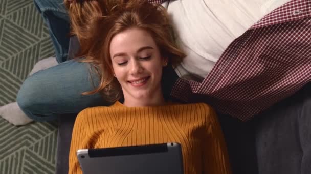 Üst görüntüde çilli bir kızın erkek arkadaşının dizlerinin üzerinde yatan tablete gülümsediği bir video. — Stok video