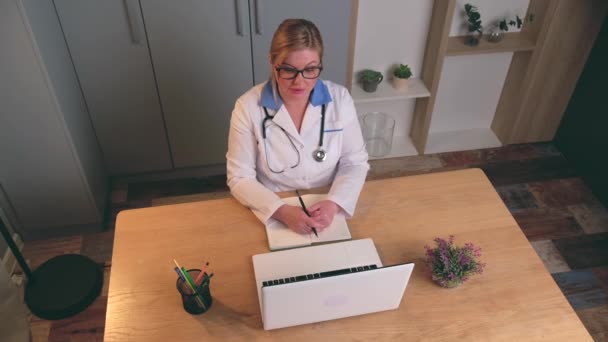 Vit blond läkare med glasögon sitter vid den bärbara datorn och har ett videomöte med en patient som förklarar nästa steg i kurprogrammet — Stockvideo