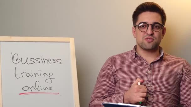 Jonge jongen met een bril is het hebben van online business training lessen spreken op de camera en glimlach in de buurt van een bord — Stockvideo