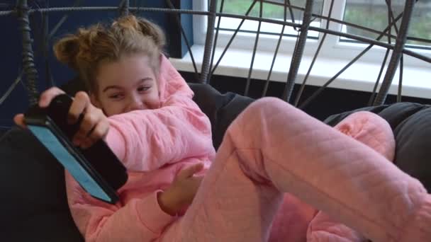 ピンクのパジャマを着たかわいい女の子が家のハンモックに横になって、近くの誰かにそれを示す電話で遊んでいると笑顔 — ストック動画