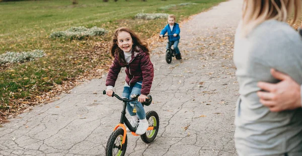 Beyaz kız ve erkek kardeşi parkta bisiklet sürerken ebeveynlerinin kucaklaşmasını izliyorlar. — Stok fotoğraf