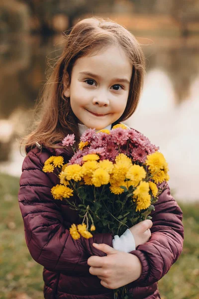 Linda menina caucasiana olhando para a câmera e posando com algumas flores com um lago no fundo no parque — Fotografia de Stock