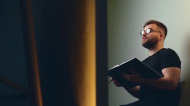 Genç sakallı öğretmen stüdyoda ders veriyor. Kameraya konuşuyor ve elinde bir defter tutuyor.