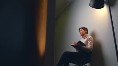 Beyaz kadın, elinde bir defter olan beyaz bir duvarda çevrimiçi dersler alırken kameraya konuşuyor.
