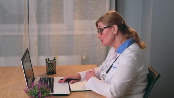Senior blonde Ärztin hat Online-Untersuchung mit einem Patienten spricht am Laptop im Büro — Stockvideo