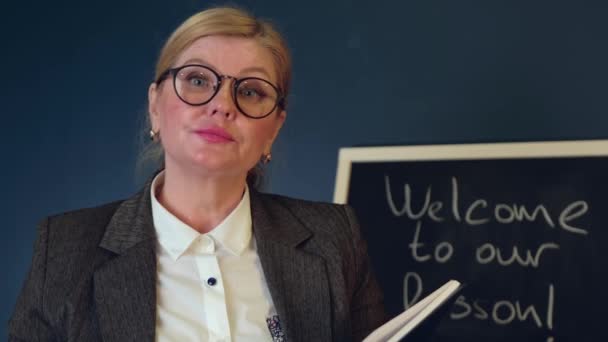 Professor sênior loira com óculos está tendo aulas on-line falando na frente de um quadro-negro — Vídeo de Stock