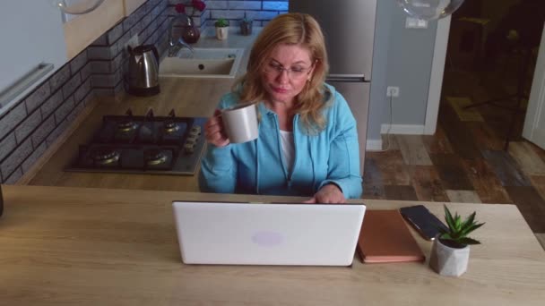 Bovenaanzicht van een drukke oudere vrouw die thee drinkt en op afstand werkt aan de computer in de keuken — Stockvideo