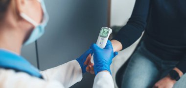 Yüzünde tıbbi maske olan beyaz bir doktor muayene sırasında hastanın vücut ısısını ölçüyor.