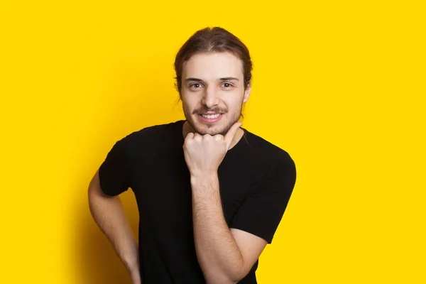 Schöner bärtiger Mann mit langen Haaren berührt ihr Kinn mit der Faust und lächelt in die Kamera an einer gelben Studiowand — Stockfoto