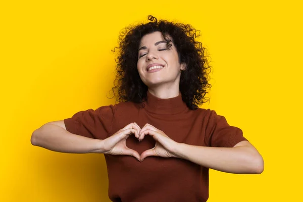 Bruneta femeie cu păr creț este gesturing semnul de dragoste cu mâinile ei zâmbind pe un perete studio galben — Fotografie, imagine de stoc