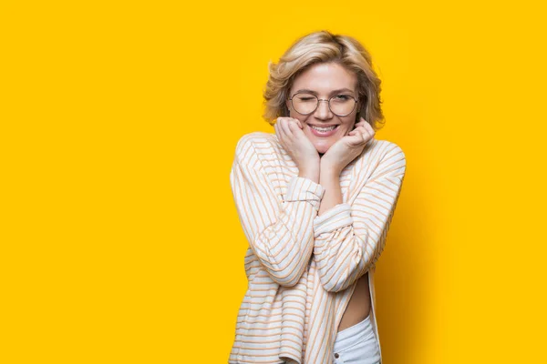 Mulher loira com óculos está anunciando algo em uma parede de estúdio amarelo com espaço livre sorrindo e tocando seu queixo — Fotografia de Stock