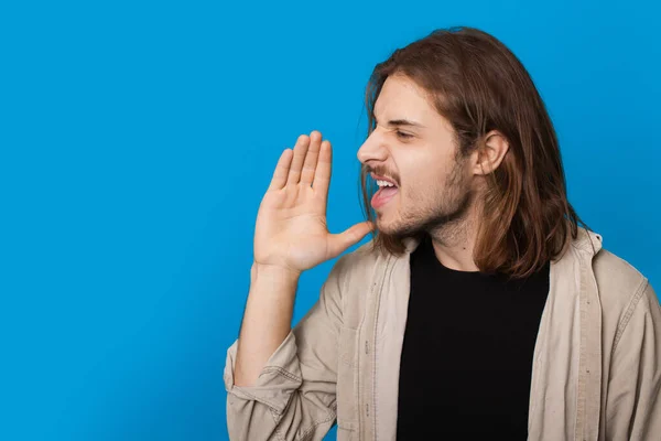 Homem caucasiano com barba e cabelos longos está gritando em uma parede de estúdio azul com espaço livre anunciando algo — Fotografia de Stock