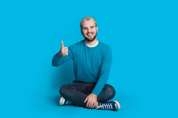 Foto monocromática de um homem caucasiano com cabelo loiro sentado no chão no estúdio posando em uma parede azul — Fotografia de Stock