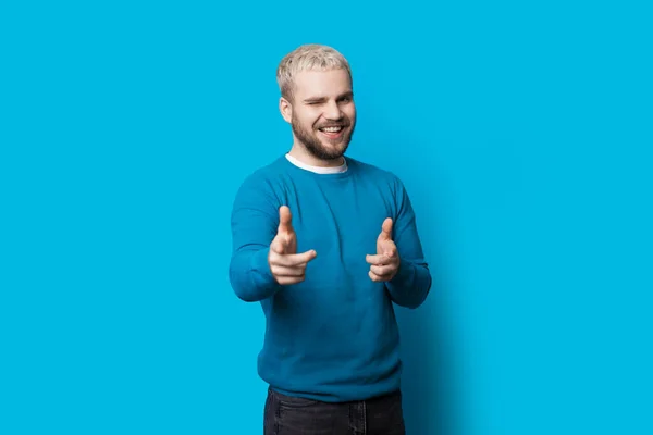 Caucasian man with blonde hair and beard is pointing at camera blinking and smile on a blue studio wall