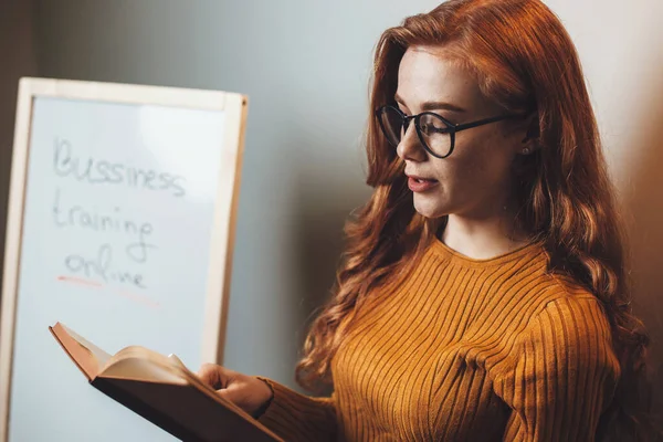Ingefära kvinna med glasögon och fräknar läser från en bok under online business classes — Stockfoto