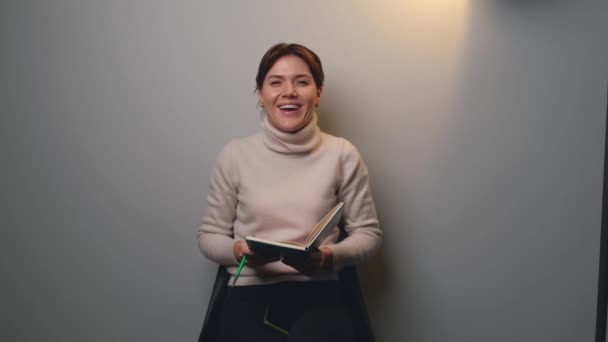 Чарівна жінка сміється з камери, тримаючи книгу на сірій стіні студії — стокове відео
