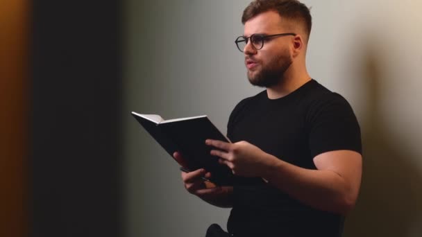Rapaz barbudo está lendo algo do livro para alguém sentado em uma parede de estúdio cinza usando óculos e camiseta preta — Vídeo de Stock