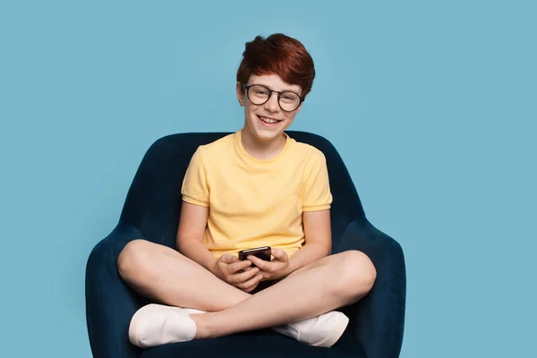 Ginger boy está usando um telefone e sorrir para a câmera embora óculos sentados em poltrona em uma parede azul estúdio — Fotografia de Stock