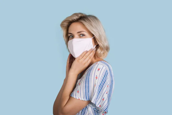 Entzückendes blondes Mädchen mit medizinischer Maske lächelt in einem Kleid an einer blauen Wand in die Kamera — Stockfoto