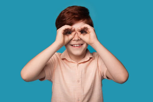 Caucasiano gengibre menino está gesticulando como ter binóculos nos olhos sorrindo em uma parede de estúdio azul — Fotografia de Stock