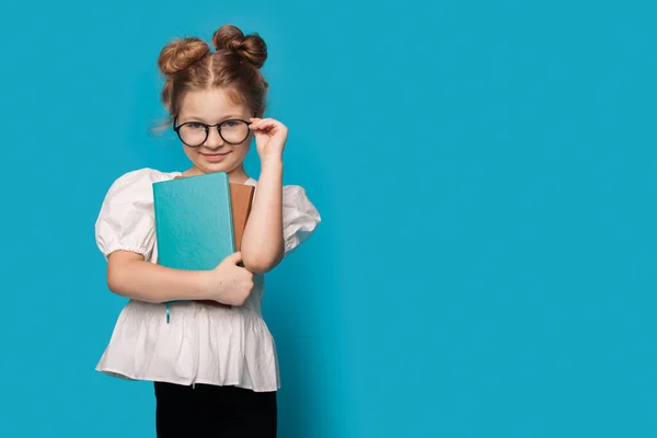 Pequeña colegiala tocando sus gafas y abrazando algunos libros en una pared azul con espacio libre — Foto de Stock