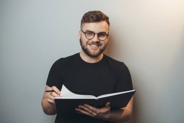 Sakallı adam elinde kitap tutarken gözlük takıyor ve gri bir stüdyonun duvarındaki kameraya gülümsüyor.