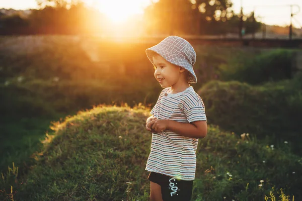 Menino encantador com chapéu azul está posando em um campo verde sorrindo ao sol — Fotografia de Stock