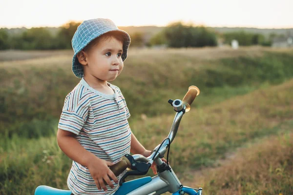 Mavi şapkalı sevimli çocuk bisiklet sürüyor, güneş ışığına karşı poz veriyor. — Stok fotoğraf