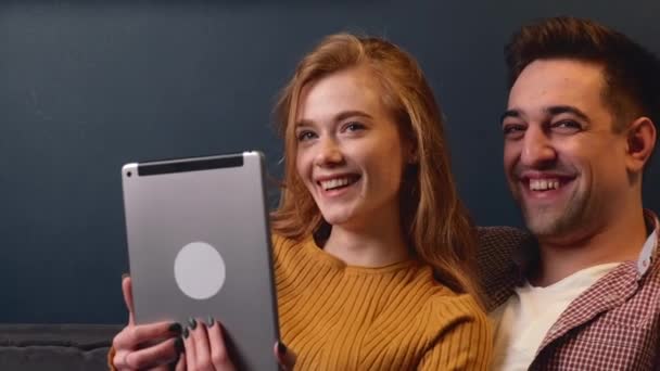 Mujer adorable con el pelo rojo y pecas sonríe mientras usa una tableta cerca de su amante en la cama — Vídeo de stock